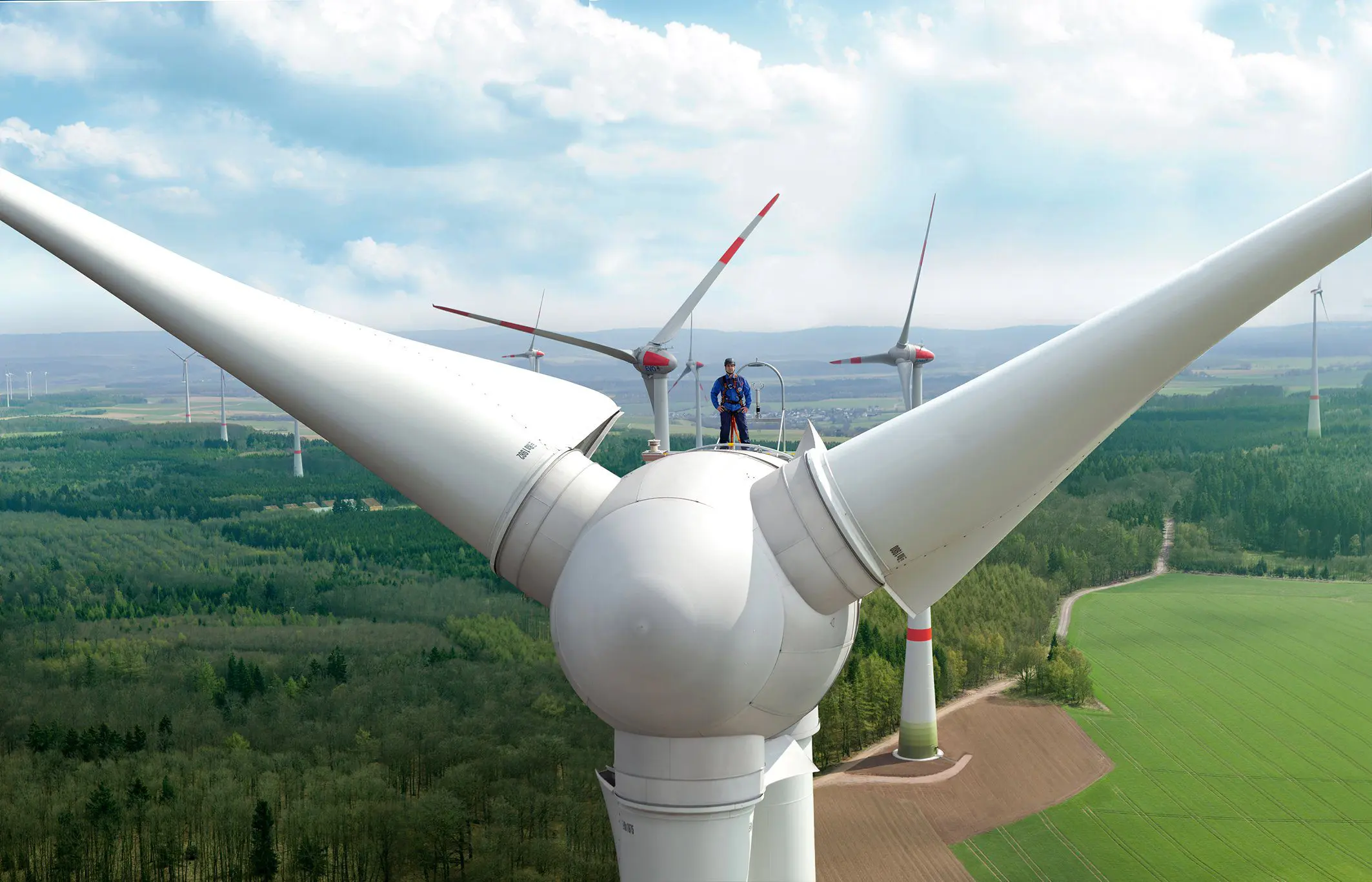 Das Bild zeigt eine große Windkraftanlage inmitten einer grünen, bewaldeten Landschaft. Auf der Anlage steht ein Techniker der MVV Energie mit Sicherheitsausrüstung, im Hintergrund erstrecken sich mehrere weitere Windräder.