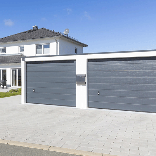 Eine graue Doppelgarage vor blauem Himmel und einem weißen Wohnhaus.