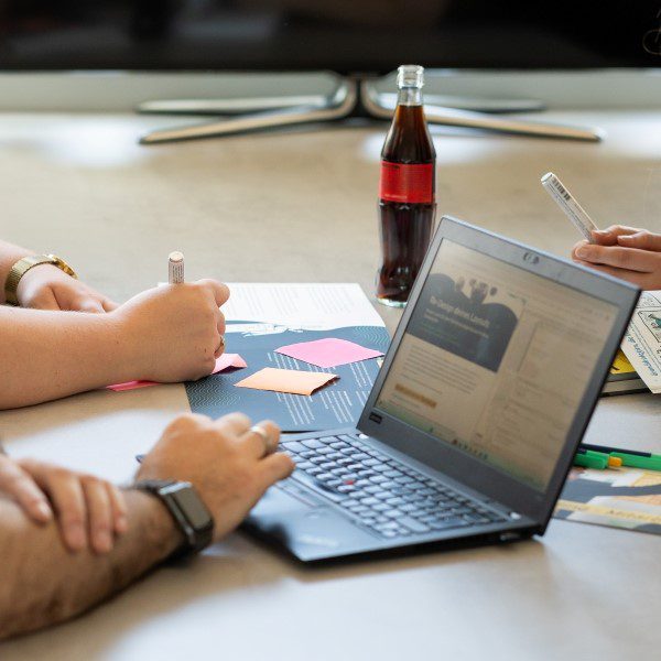 Ein Foto zeigt einen Laptop mittig auf einem Tisch. Um den Laptop sitzen mehrere Personen, deren Hände zu sehen sind und am Laptop arbeiten und Notizen machen. Im Hintergrund steht eine Getränkeflasche.