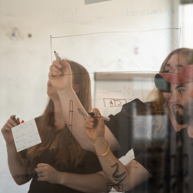 Ein Foto zeigt unsere Mitarbeitenden Saskia, Nina-Marie und Simi aus dem Kreationsteam, die hinter einer Glasscheibe stehen und gemeinsam etwas an die Wand schreiben.